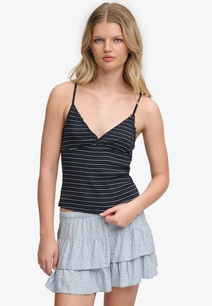 Jeune femme aux longs cheveux blonds portant un caraco rayé noir et blanc et un short bleu clair à volants superposés.