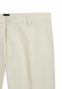 Beige katoenen chinos met een recht gesneden ontwerp, zijzakken en een knoopsluiting. De stof heeft een gladde, iets textuurachtige afwerking.