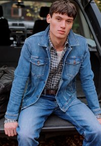 Jachetă din denim de culoare bleu deschis, cu două buzunare pe piept, purtată peste o cămașă cu carouri. Blugi asortați cu jacheta, cu un curea maro. O geantă maro în fundal.
