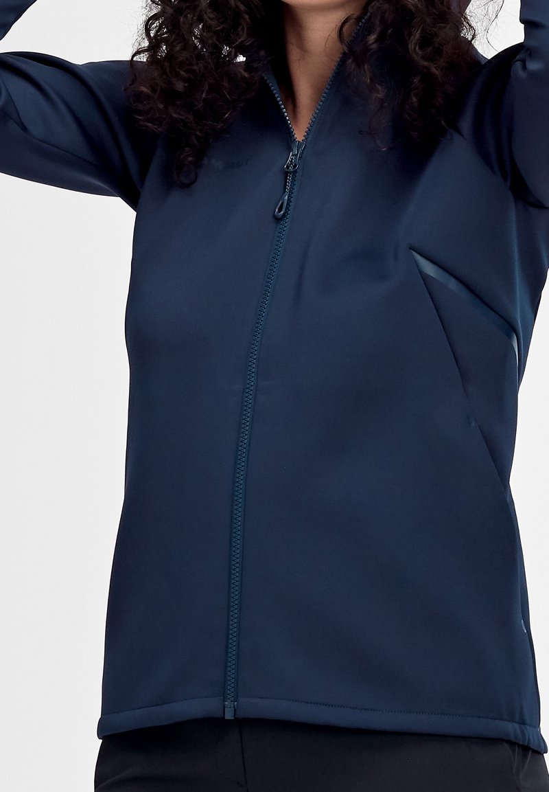 Femme portant une veste zippée bleu marine à manches longues, visible des épaules aux hanches sur un fond uni.