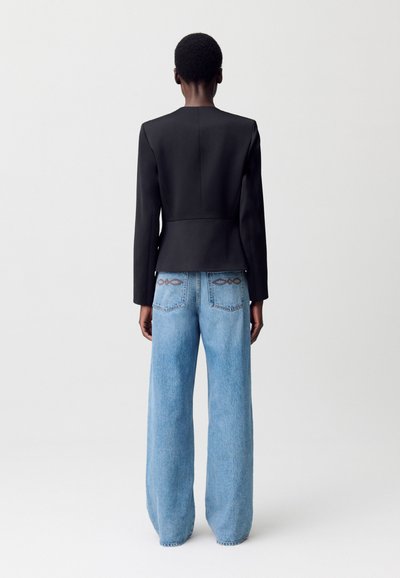 Una persona con el pelo corto lleva una chaqueta negra ajustada y pantalones vaqueros de pierna ancha, de color azul claro, vista desde atrás contra un fondo minimalista.