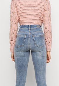 Suéter de malha rosa claro com um padrão texturizado, combinado com jeans azuis de cintura alta com bolsos traseiros e um efeito lavado.