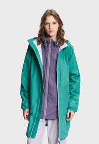 Jeune femme portant un long imperméable bleu sarcelle par-dessus une veste zippée violette et un pantalon de couleur claire, debout devant un fond clair uni.