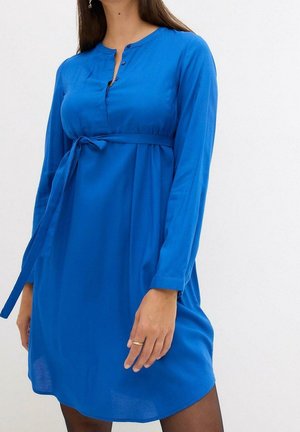 Vrouw die een koningsblauwe jurk met lange mouwen draagt, met een strikceintuur in de taille en knopen bij de hals, staand tegen een effen witte achtergrond.