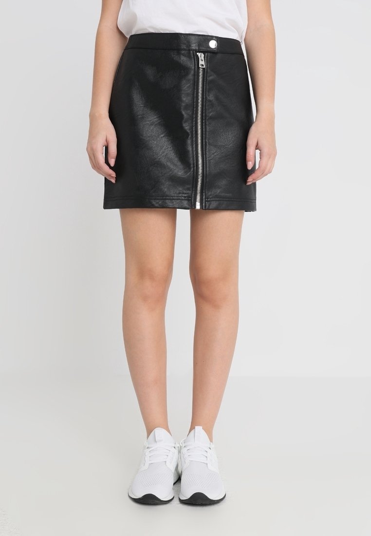 Femme portant une mini jupe en simili cuir noir avec une fermeture éclair devant et des baskets blanches, debout devant un fond uni.