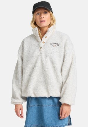 Pull en polaire blanc et duveteux avec un col haut, trois boutons en bois et un logo. Associé à une jupe en denim et une casquette noire.