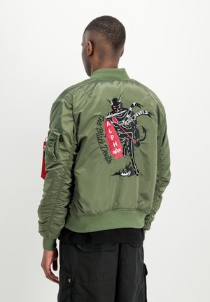 Man met de rug naar de camera draagt een groene bomberjack met een geborduurde zwarte duivel die een rood skateboard vasthoudt met het label "ALPHA" op de rug.