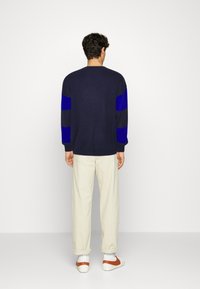 Pull bleu marine avec des rayures bleues et noires sur les manches, associé à un pantalon beige clair et des baskets blanches avec des accents orange.