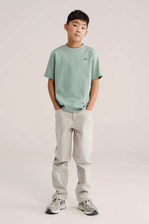 Fiú bézs nadrág zsebében tartott kézzel, világoszöld rövid ujjú inget és szürke sportcipőt viselve, egyszínű háttér előtt.
