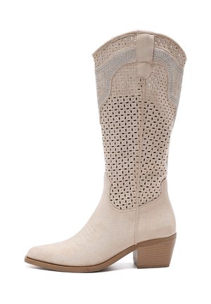 Botte western en daim beige avec un bout pointu, une tige perforée et un talon empilé. Couture décorative notable et accents texturés.