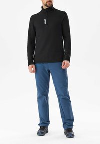 Schwarzer Zip-Fleece-Pullover mit langen Ärmeln, kombiniert mit blauen Hosen mit Taschen. Das Model trägt schwarze Wanderschuhe. Minimalistisches Design.