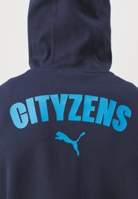 Mikina s kapucňou v námorníckej modrej farbe s výrazným tyrkysovým textom "CITYZENS" a malým bielym logom Puma, s jemnou textúrou a klokankovou kapsou.