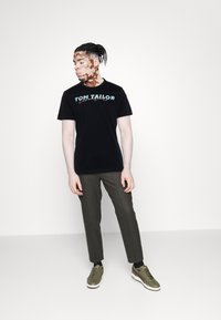 T-shirt noir à manches courtes avec "TOM TAILOR" en lettres teal, associé à un pantalon vert olive et des baskets basses avec des semelles blanches.