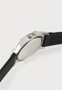 Reloj de metal plateado con caja redonda, correa de goma negra texturizada y corona lateral. La parte trasera del reloj es lisa y está etiquetada.