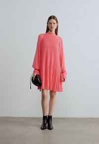 Abito rosa plissettato con maniche lunghe e collo alto. Indossato con stivaletti neri e una piccola borsa nera. Tessuto liscio, design fluido.