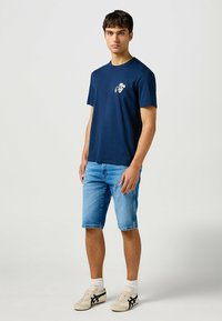 Camisa de algodón azul marino con un pequeño gráfico en el pecho, combinada con pantalones cortos de mezclilla claros y zapatillas beige con acentos negros.