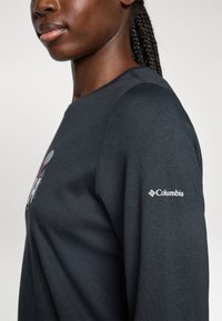 Detailný záber osoby v tmavom tričku s dlhým rukávom, na ktorom je logo Columbia na rukáve a čiastočne viditeľná grafika na hrudi.