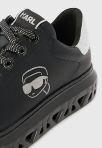 Black sneaker with smooth synthetic upper, reflective silver accents, patterned sole, and logo detail featuring sunglasses on the side.