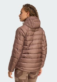 Smeđa puffy jakna s kapuljačom, izrađena od sjajnog materijala. Ima horizontalne šavne detalje i prilagođen dizajn, nosi se preko uzorčanih tajica.