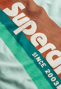 Light blue t-shirt with textured white "superdry" text, featuring horizontal stripes in teal and brown, labeled "SINCE 2003."