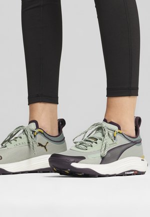 Zapatillas deportivas verde claro con tejido texturizado, acentos en púrpura oscuro y suelas grises. Incluyen cordones y marca en el lateral.