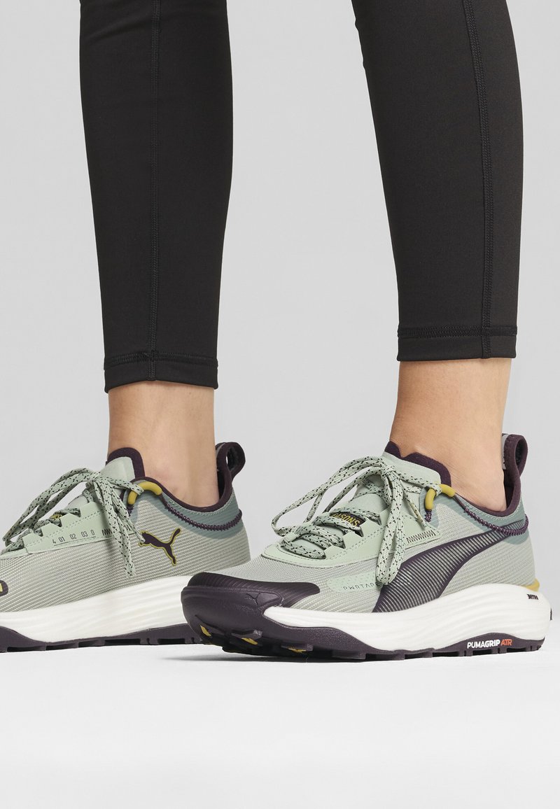 Light green athletic shoes with textured fabric, dark purple accents, and grey soles. Features include laces and branding on the side.