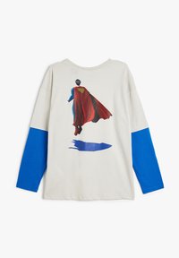 Cremefarbenes Langarm-T-Shirt mit blauen Akzenten und einem bedruckten Design eines Charakters in einem roten Umhang, der einen Schatten darunter hat.