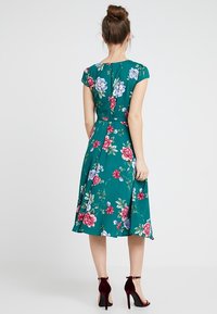 Květinové šaty s zelenou látkou, krátkými rukávy, vypasovaným pasem a sukní ke kolenům s velkými červenými, růžovými a modrými květy.