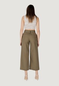 Pantaloni larghi color khaki con vita alta e tasche posteriori. La modella indossa un top corto in pizzo bianco e tacchi trasparenti.