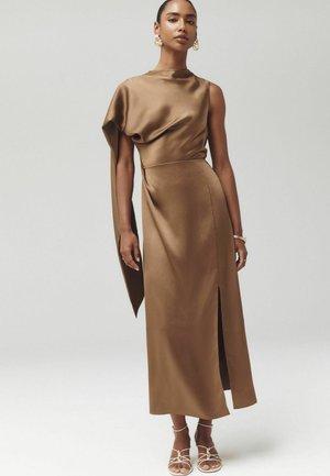 Woman wearing a sleeveless brown satin dress with asymmetric drape and front slit, accessorized with gold earrings, bracelet, and white strappy heels.