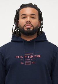 Felpa blu navy in cotone, con un logo frontale in testo rosso, cordini e tasca a marsupio. Design semplice con una vestibilità casual.