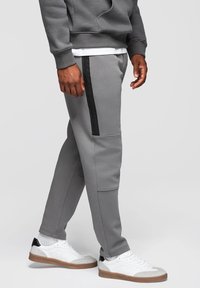 Szare dresowe spodnie z czarnym paskiem bocznym, wykonane z miękkiego materiału. Projekt charakteryzuje się dopasowanym krojem oraz bocznymi kieszeniami, noszone z białymi sneakersami.
