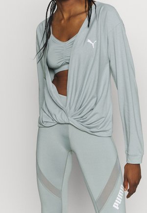 Vrouw draagt een lichtgrijs Puma sportoutfit bestaande uit een jack met gedraaide zoom, een gerimpelde sportbeha en legging met mesh panelen en logo.