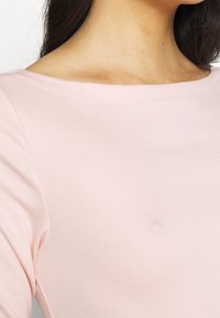 Helles rosa Langarmshirt mit Rundhalsausschnitt. Der Stoff ist glatt und dehnbar, mit gerippten Nähten entlang des Ausschnitts und der Ärmel.