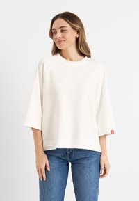 Lichtcrème oversized sweatshirt met korte raglanmouwen, minimalistisch ontwerp en klein logo op de borst. Gecombineerd met blauwe spijkerbroek.