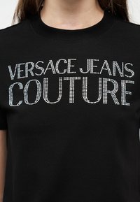 Μαύρο μπλουζάκι με την επιγραφή «VERSACE JEANS COUTURE» σε λευκά γράμματα με κουκκίδες, φορεμένο από ένα άτομο με μακριά μαλλιά και ανοιχτόχρωμο δέρμα.