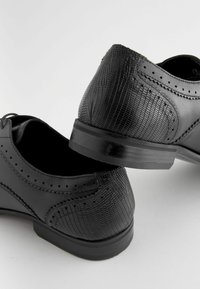 Mustad mustrinimed, millel on sile nahk, tekstuuritud reljeefmustriga kontsadel ja brogue detailid külgedel.