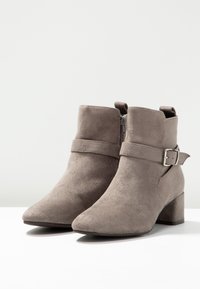Par de botines de ante gris con punta afilada, tacones de bloque, cremallera lateral y correa decorativa con hebilla sobre una superficie blanca.