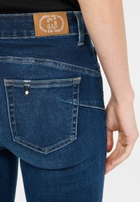Los pantalones vaqueros de mezclilla azul oscuro presentan un parche de cuero marrón con detalles metálicos en la cintura, y un pequeño acento de hardware plateado en el bolsillo trasero.