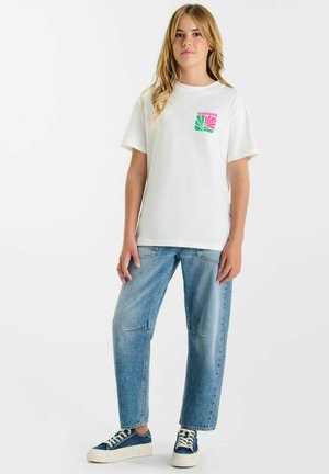 Jeune femme debout portant un T-shirt blanc avec un graphisme rose-vert, un jean bleu ample et des baskets bleues sur fond blanc.