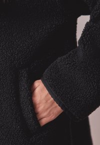 Manteau en polaire noire avec tissu texturé. Comprend une poche visible et un bord de manche contrastant noir. Apparence douce et chaude.