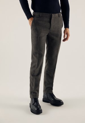 Man die donkergrijze slim-fit broek draagt, gecombineerd met zwarte leren veterschoenen en een zwarte trui met lange mouwen, staand met een hand in de zak.