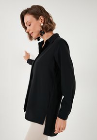 Black long-sleeve shirt with a collar and side slits. Smooth fabric, slightly loose fit, featuring minimalistic design and clean lines.