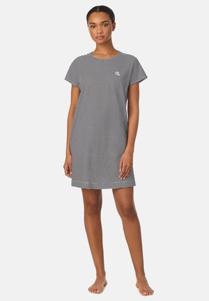 Vrouw staat op blote voeten en draagt een knielange jurk met korte mouwen, zwart-wit horizontale strepen en een klein logo op de borst.