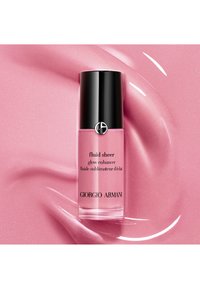 Klar glassflaske med svart hette, som inneholder en skimrende rosa væske. Etiketten sier "fluid sheer glow enhancer" og "Giorgio Armani." Myk rosa bakgrunn.