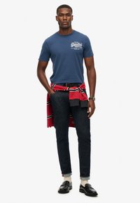 Blå bomulds T-shirt med hvid grafisk tryk, mørke denim jeans, rød- og sortstribet sweatshirt bundet om livet, og sorte loafers.