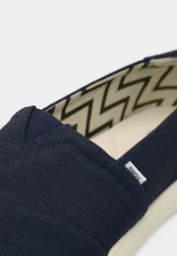 Chaussure en toile bleu marine slip-on avec semelle blanche et doublure intérieure beige à motif zigzag, avec une petite étiquette de marque en tissu sur le côté.