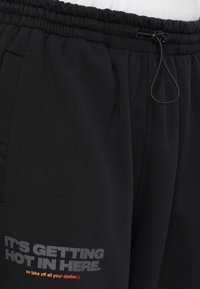 Μαύρες φόρμες με ελαστική μέση και κορδόνι. Διαθέτουν εκτυπωμένο κείμενο σε γκρι και πορτοκαλί στο αριστερό μηρό. Λείο υλικό.