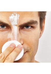 Eine Nahaufnahme einer Person, die ein Wattepad auf ihr Gesicht aufträgt, das vertikal eine weiße Creme oder Maske auf der Stirn hat.