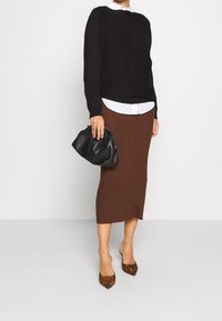 Femme portant un pull noir sur une chemise blanche, une jupe crayon midi marron, des talons imprimés léopard, tenant une pochette en cuir noir.